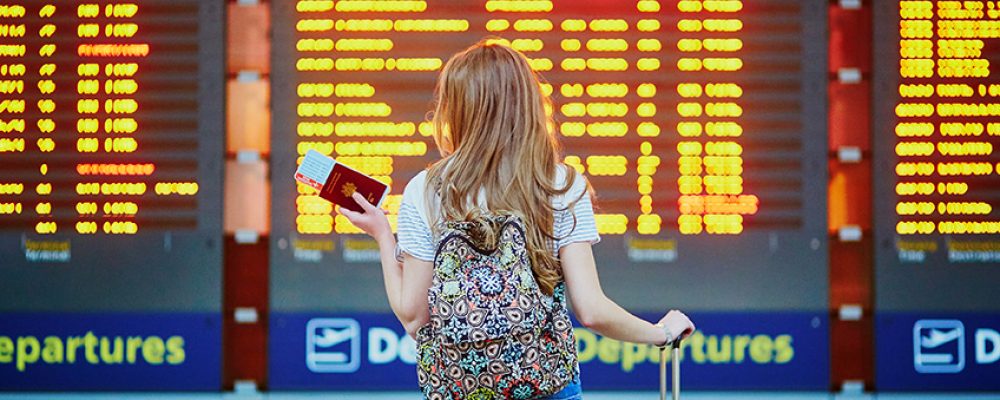 Beautiful young tourist girl with backpack and carry on luggage in international airport near flight information board
